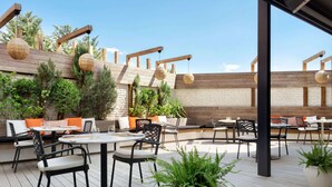 Ein Außenbereich mit einer Terrasse, runden Tischen und Stühlen, Holzbalken und kugelförmigen Hängeleuchten.