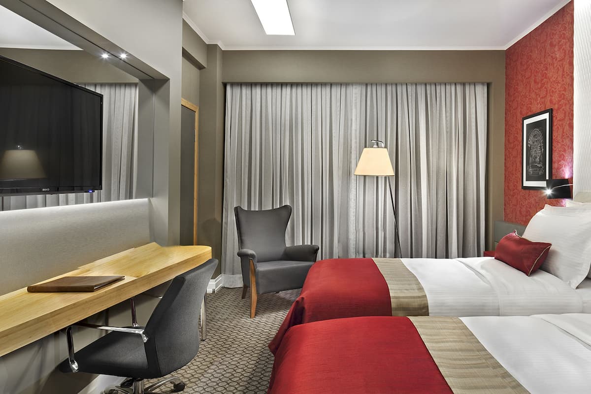 Une chambre d’hôtel avec deux lits, un bureau, une chaise, une lampe et une grande fenêtre avec des rideaux.