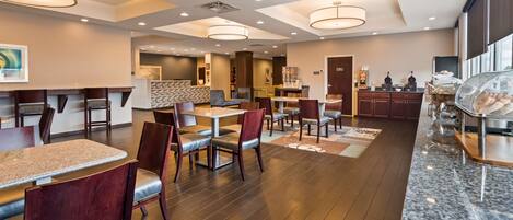 A hotel dining area with multiple tables and chairs, a kitchen in the background, and a buffet line with covered dishes.