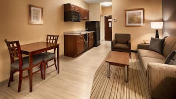 Una habitación de hotel moderna con una mesa de comedor, sillas, un sofá, una mesa de centro, una zona de cocina con refrigerador y microondas, y un cuadro en la pared.