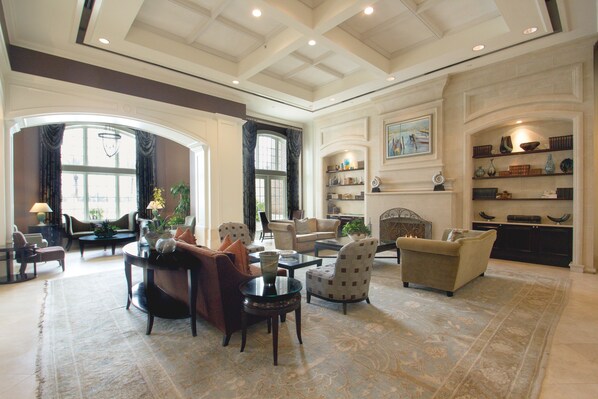 Un salon spacieux avec un plafond haut, de grandes fenêtres et une cheminée. On y trouve plusieurs coins salon avec des canapés et des fauteuils.