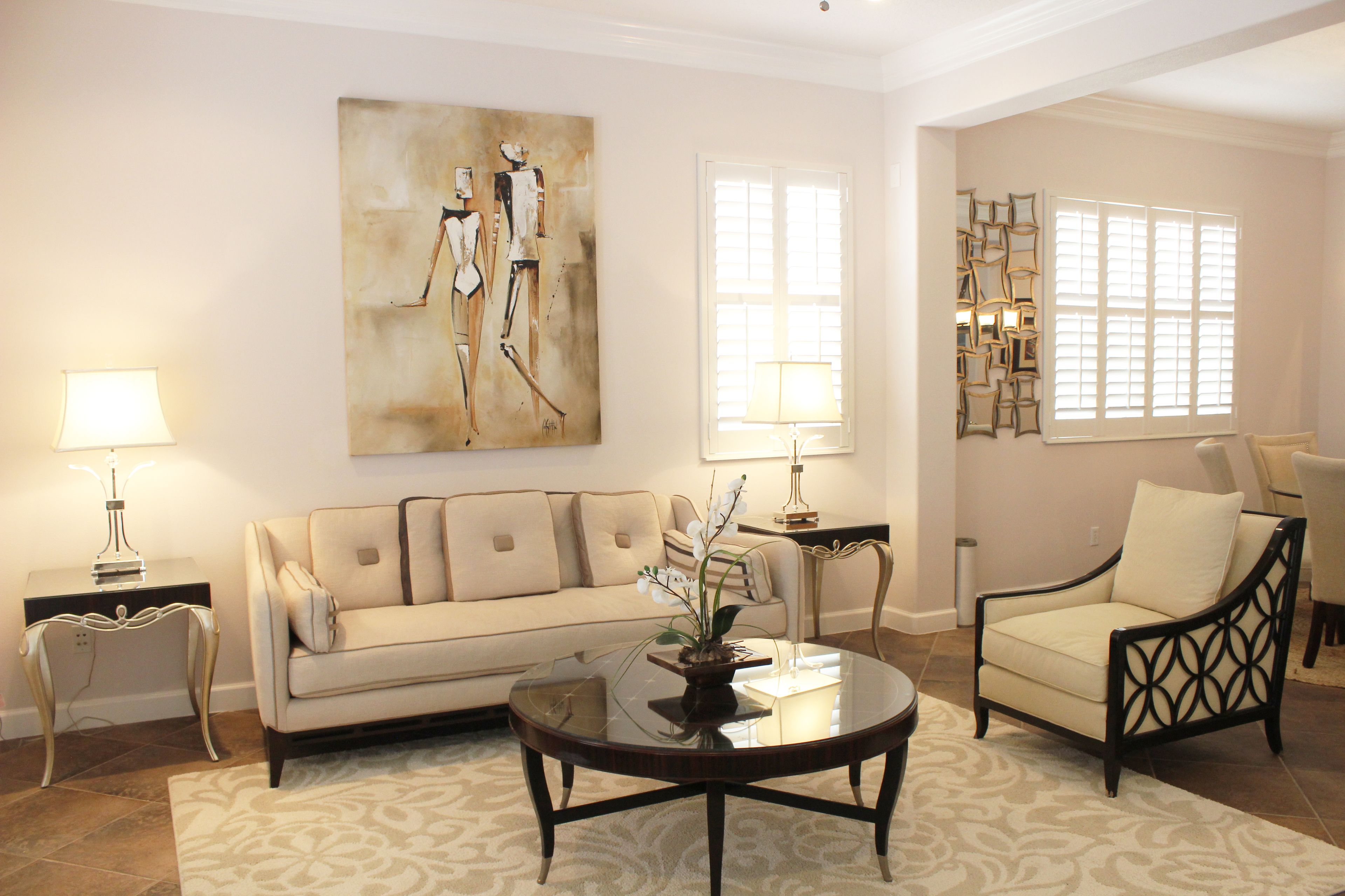 A living room with a beige sofa, a round coffee table, and a chair. There is a painting on the wall and a window with blinds.
