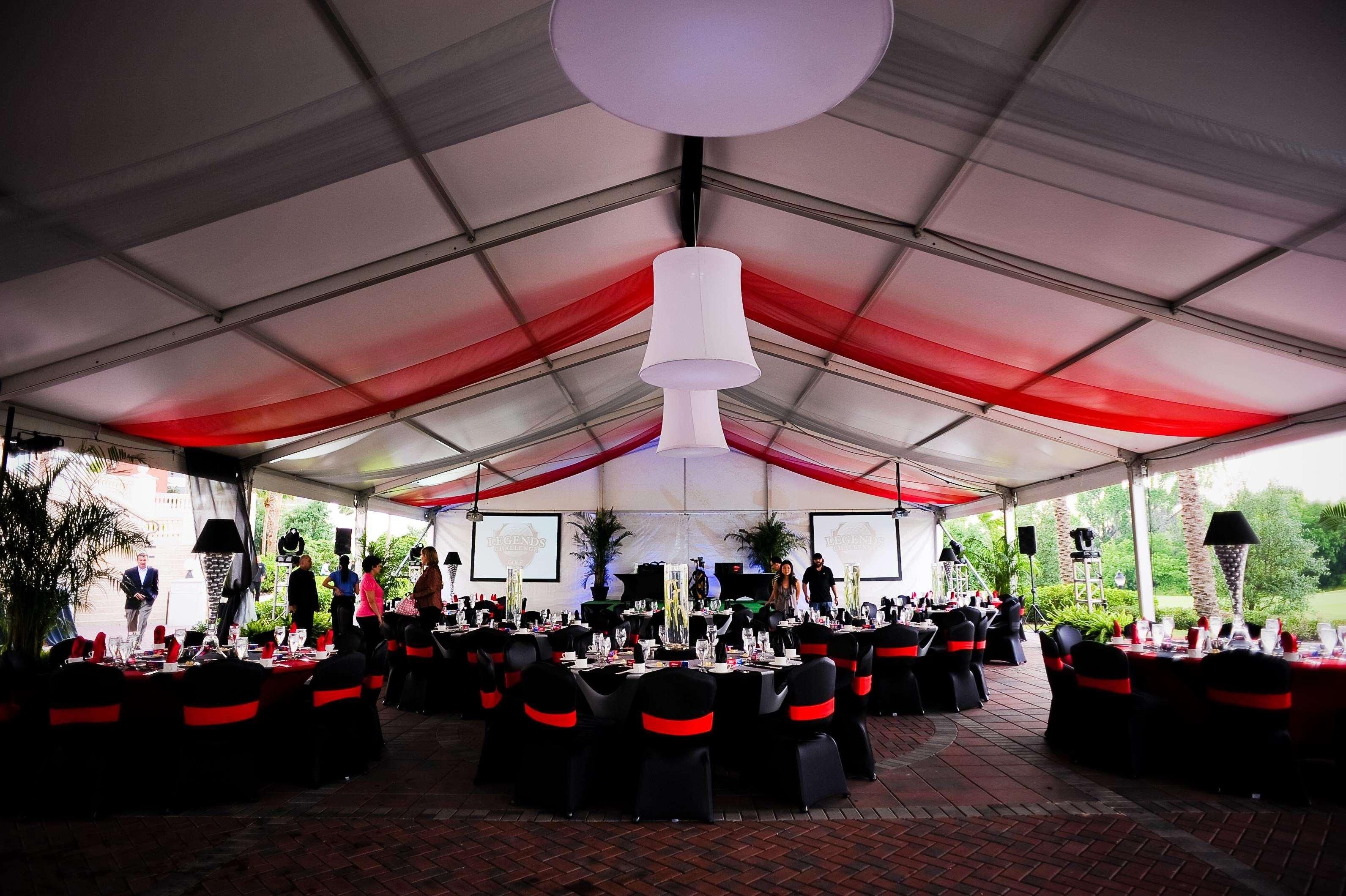 A tented outdoor event space with tables set for dining, surrounded by chairs with red sashes, and a large white hanging light fixture.
