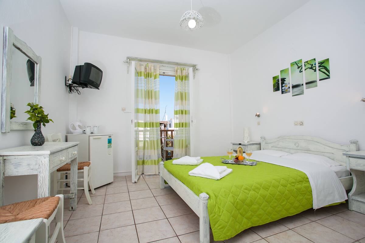 Un dormitorio con cama, escritorio, silla, refrigerador pequeño, televisor y ventana con cortinas.