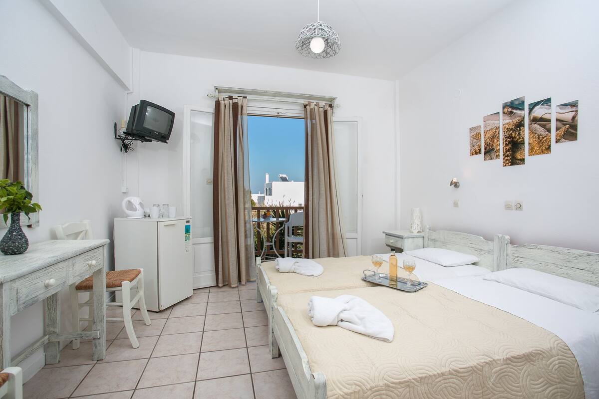 Una habitación de hotel con cama, escritorio, televisión y balcón con cortinas.