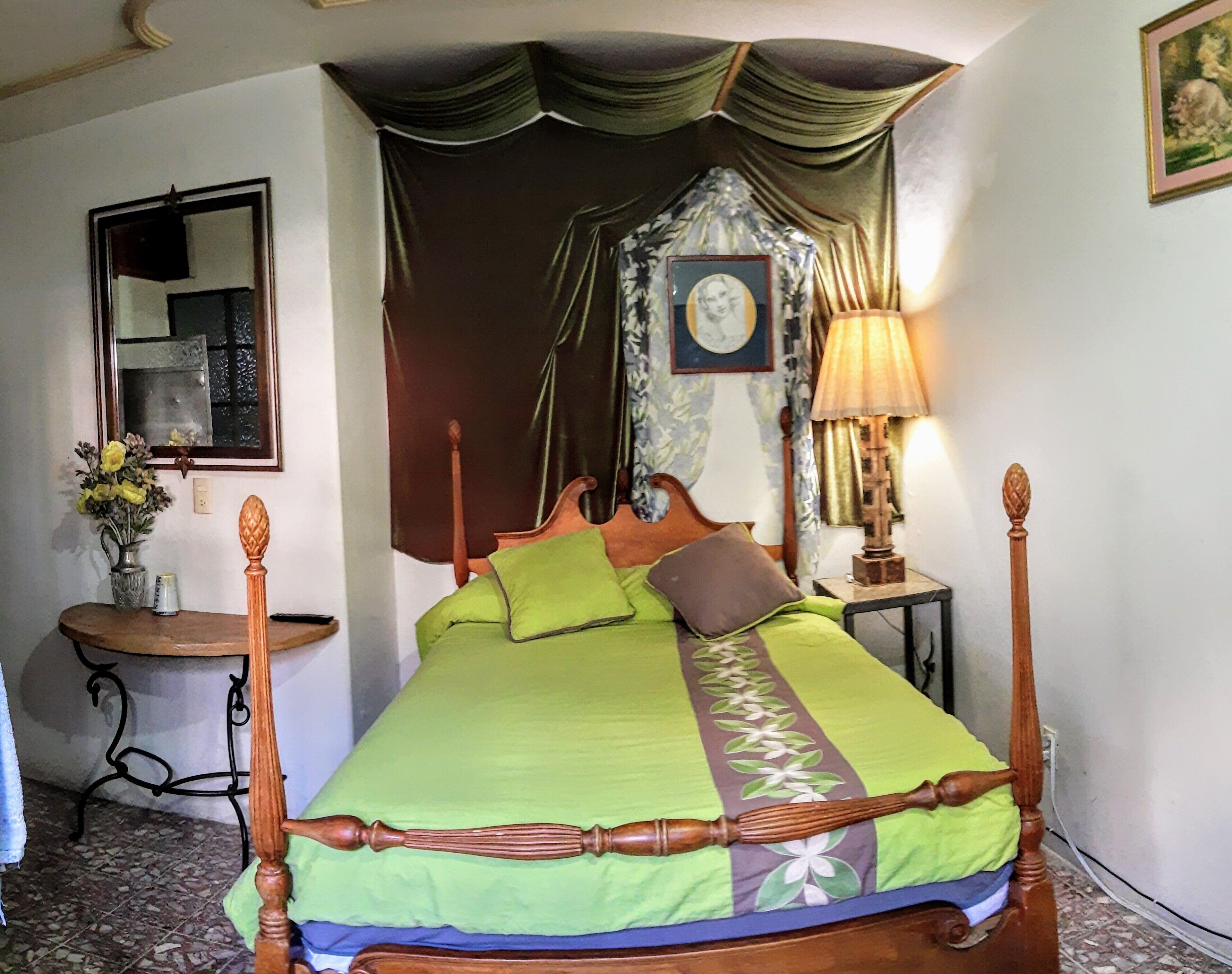 Una cama con dosel tradicional, con ropa de cama verde y marrón, una mesita de noche de madera con una lámpara, un espejo y un jarrón con flores a la izquierda.