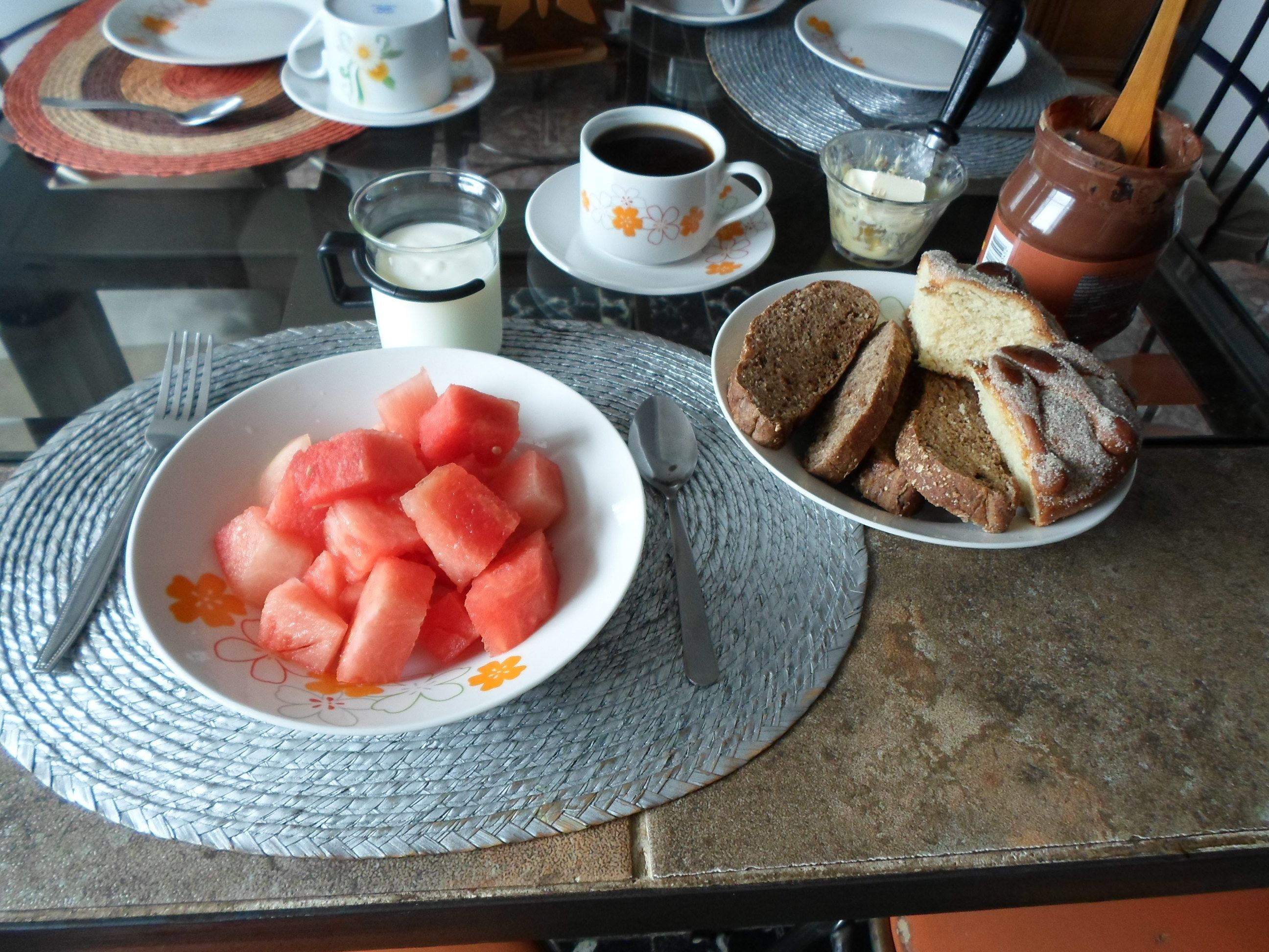 A table with a bowl of watermelon, slices of bread, a cup of coffee, and a glass of milk.