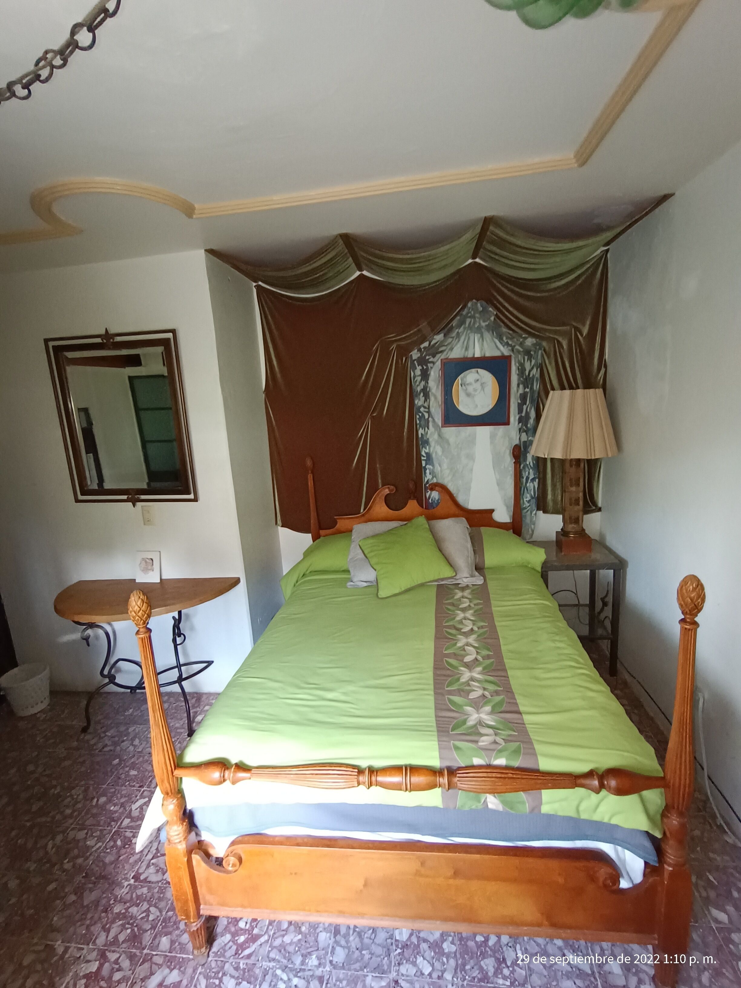 A bedroom with a wooden bed, green bedding, a mirror, a small table, and a lamp.