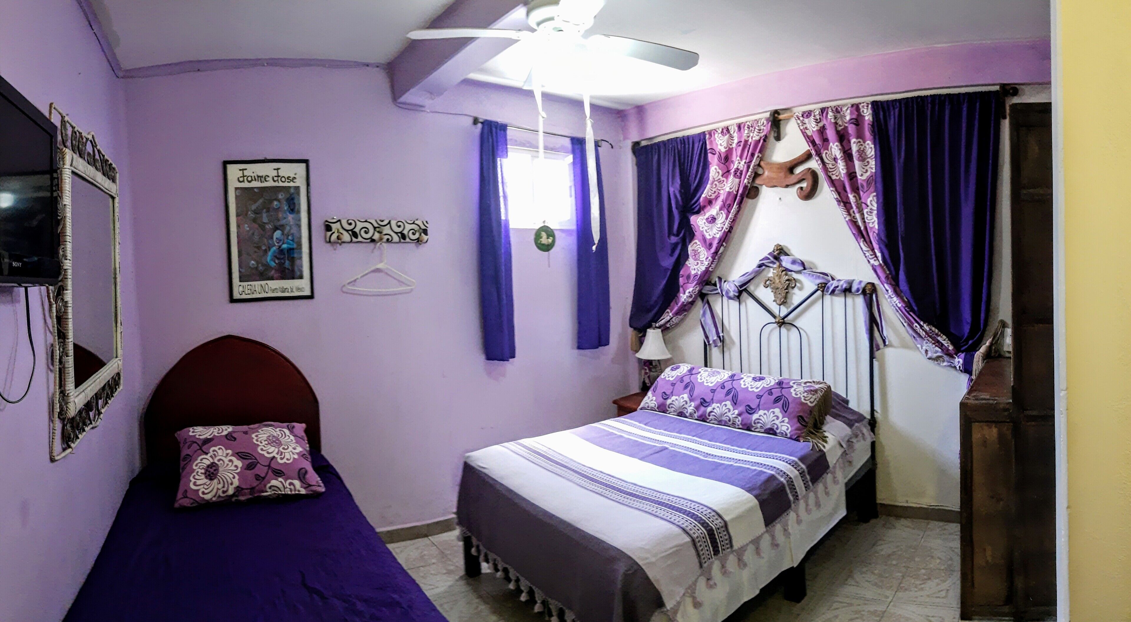 A bedroom with a bed, a bedside table, a hanging light fixture, a window with curtains, and a wall-mounted TV.