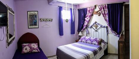 A bedroom with a bed, a bedside table, a hanging light fixture, a window with curtains, and a wall-mounted TV.