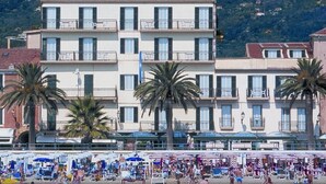 Un hôtel situé sur la côte, doté de nombreux balcons, d’une plage aménagée avec des parasols et de palmiers.