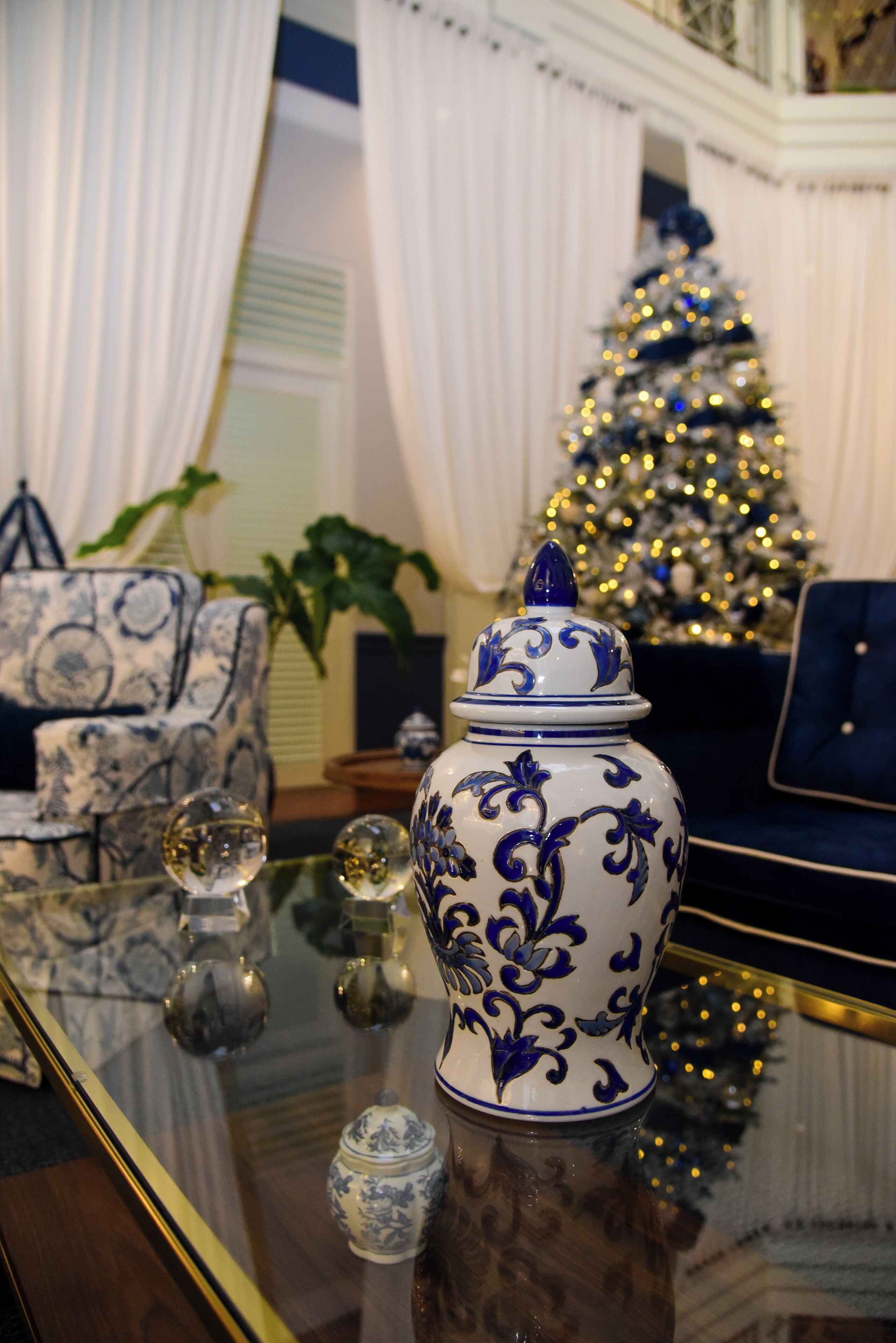Una habitación con una mesa de vidrio mostrando objetos decorativos, una silla de patrón azul y blanco, y un árbol de Navidad al fondo.