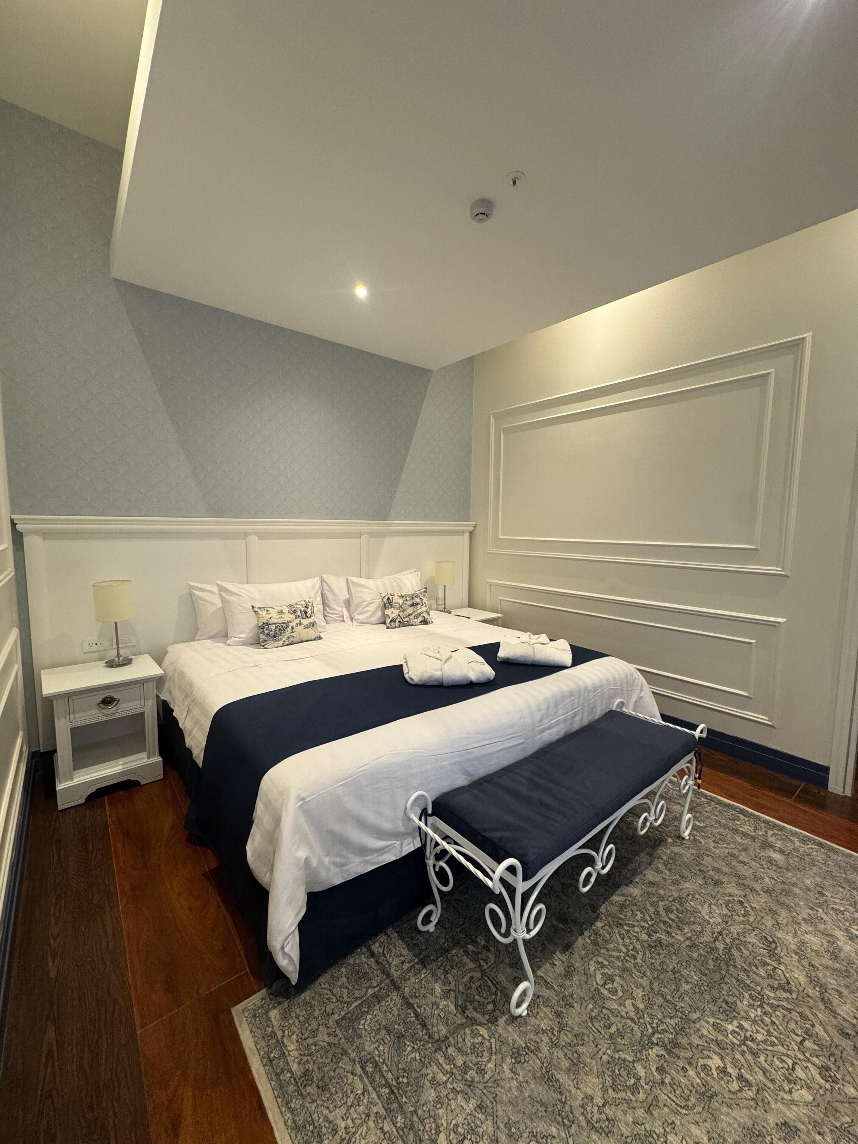 A hotel room with a large bed, bedside tables, a bench, and a patterned rug.
