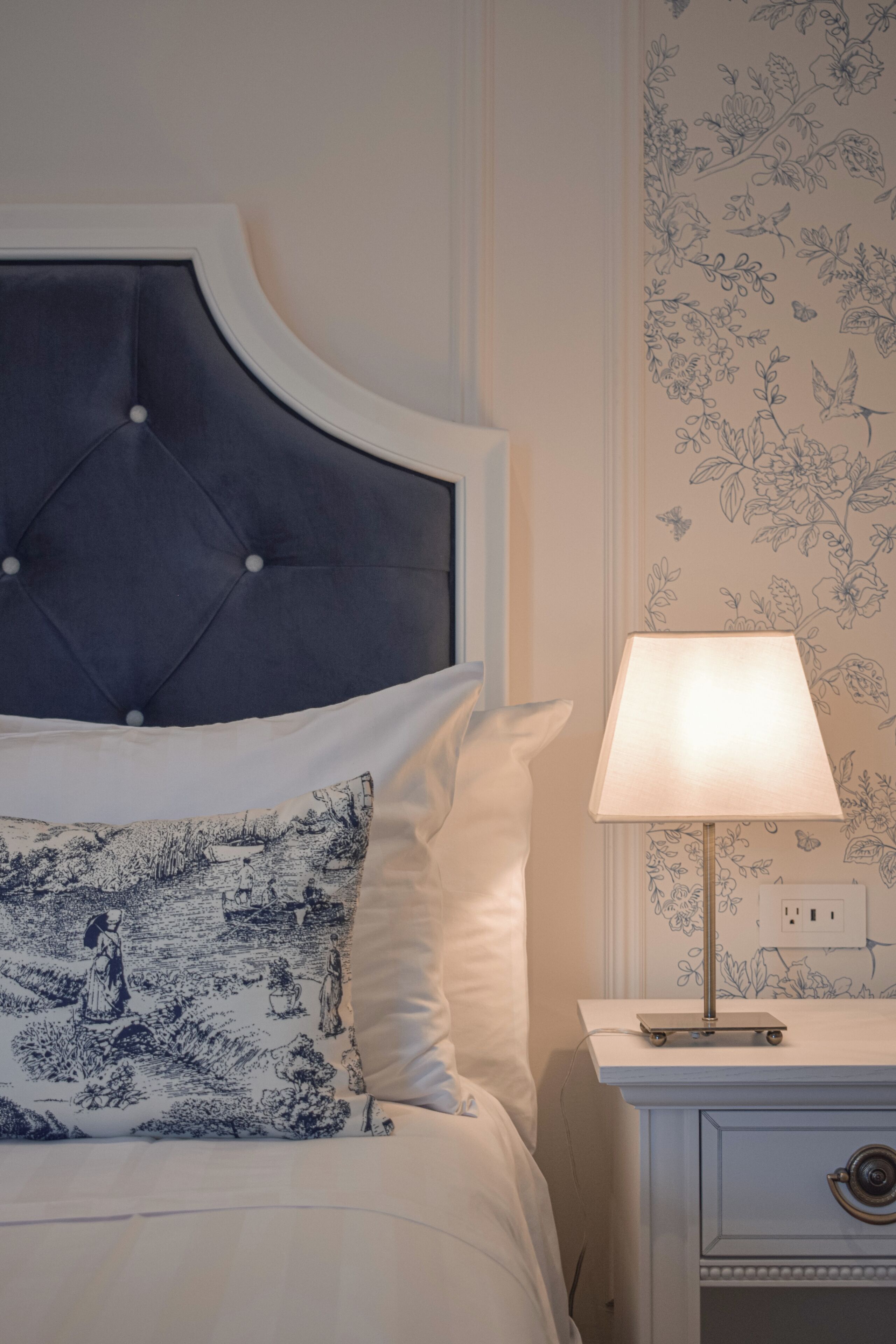 A bed with a tufted headboard, a bedside table with a lamp, and floral wallpaper.