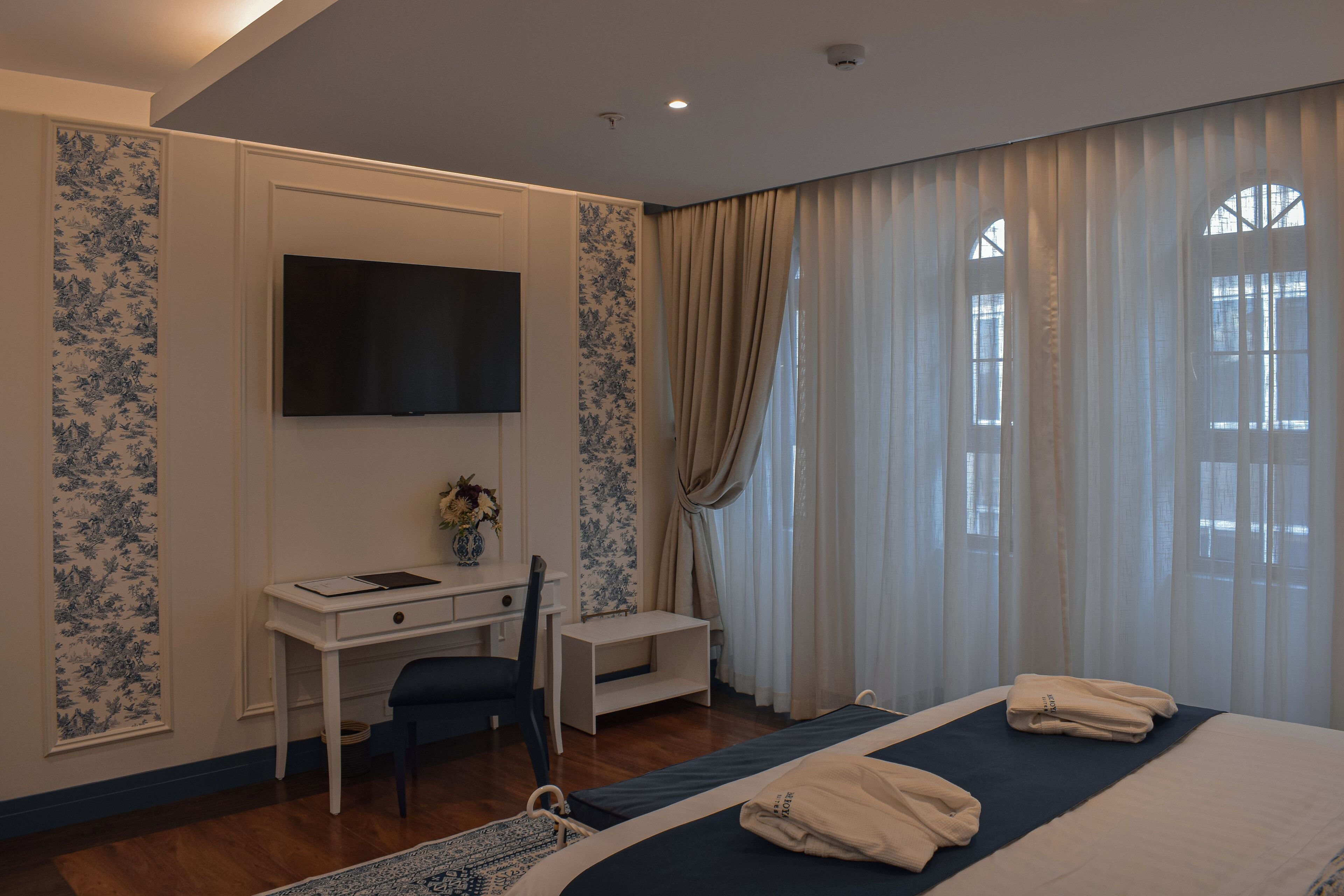 Habitación de hotel con cama, escritorio, silla, televisor y ventana con cortinas.