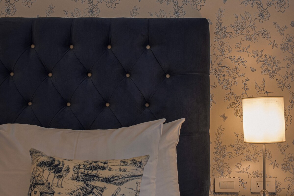A bedroom with a tufted headboard, a white bedspread, a decorative pillow, a bedside table with a lamp, and floral wallpaper.
