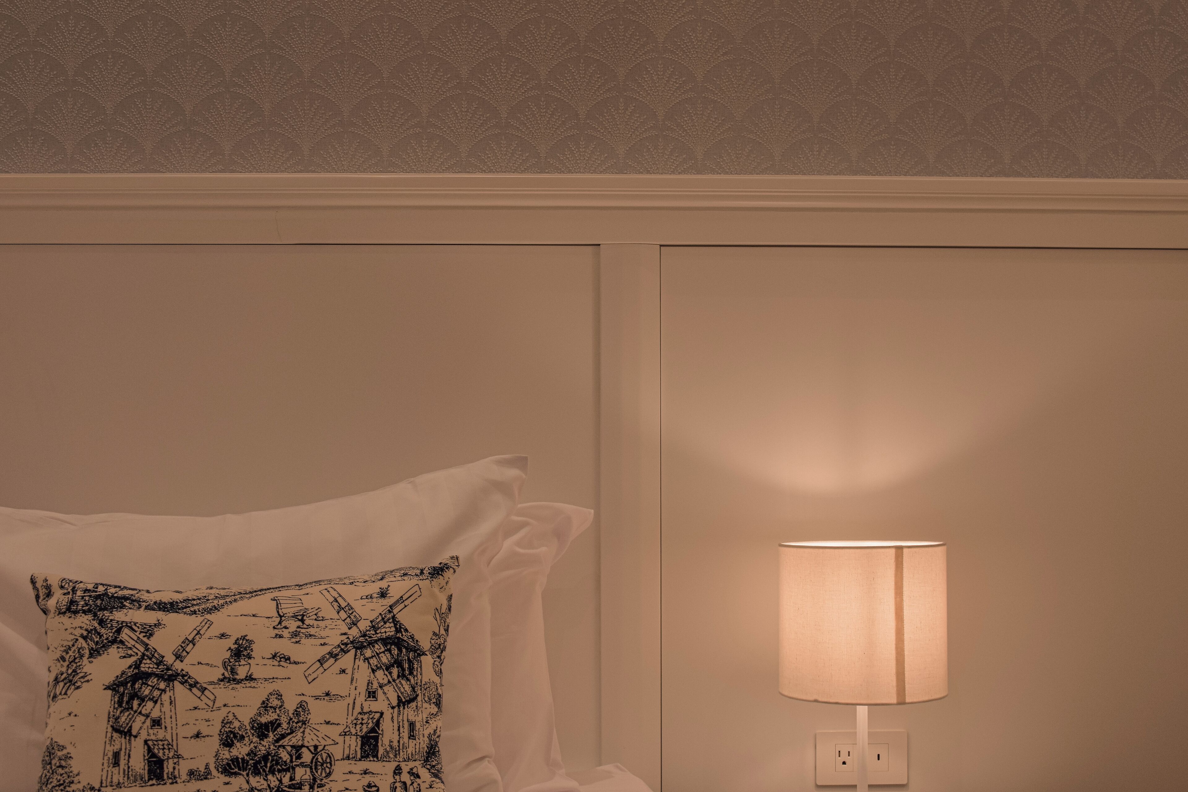 A bedroom with a patterned wall, a white pillow with a windmill design, and a bedside lamp.