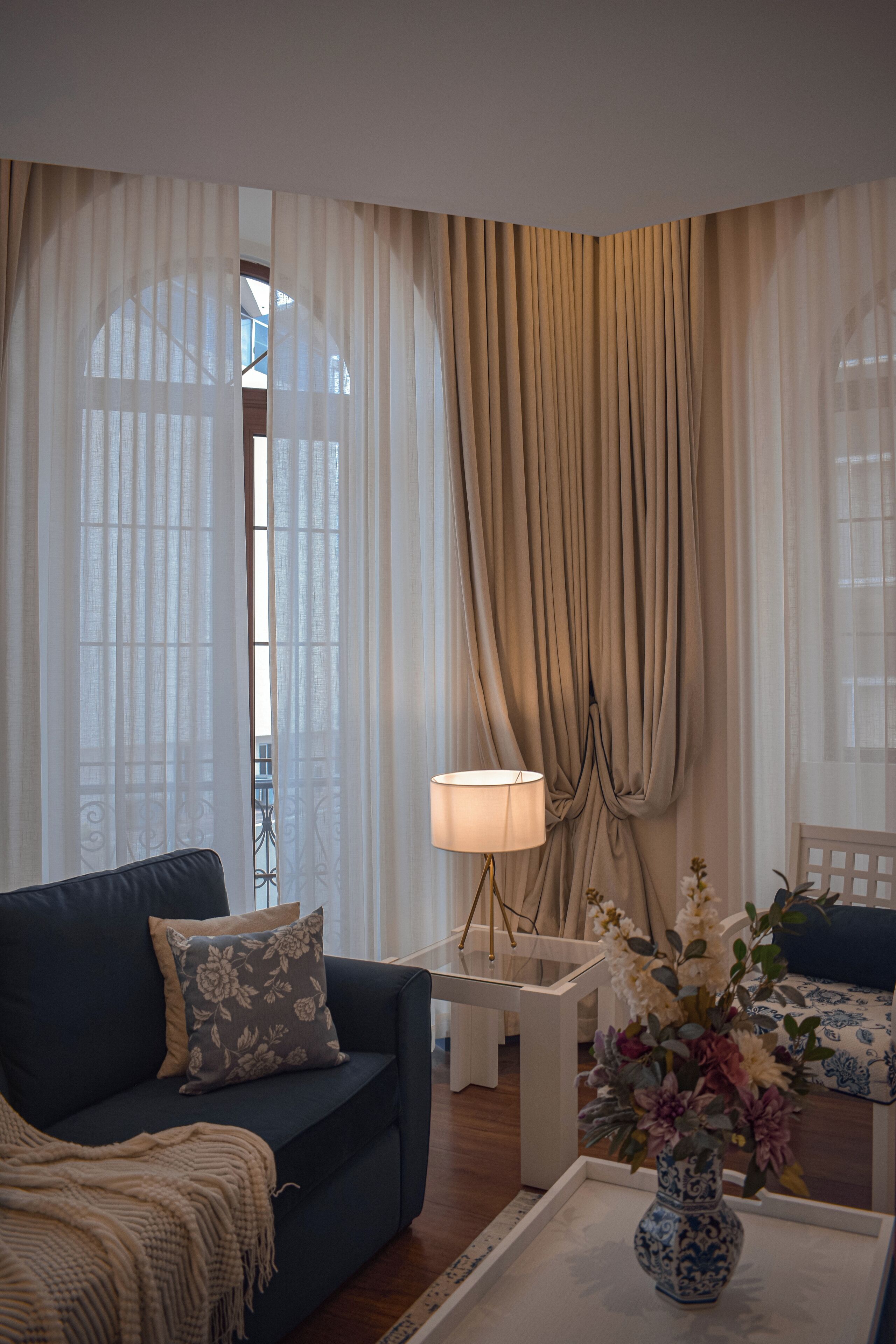 Una sala de estar con un sofá azul oscuro, una mesita auxiliar blanca, una lámpara y un jarrón con flores.