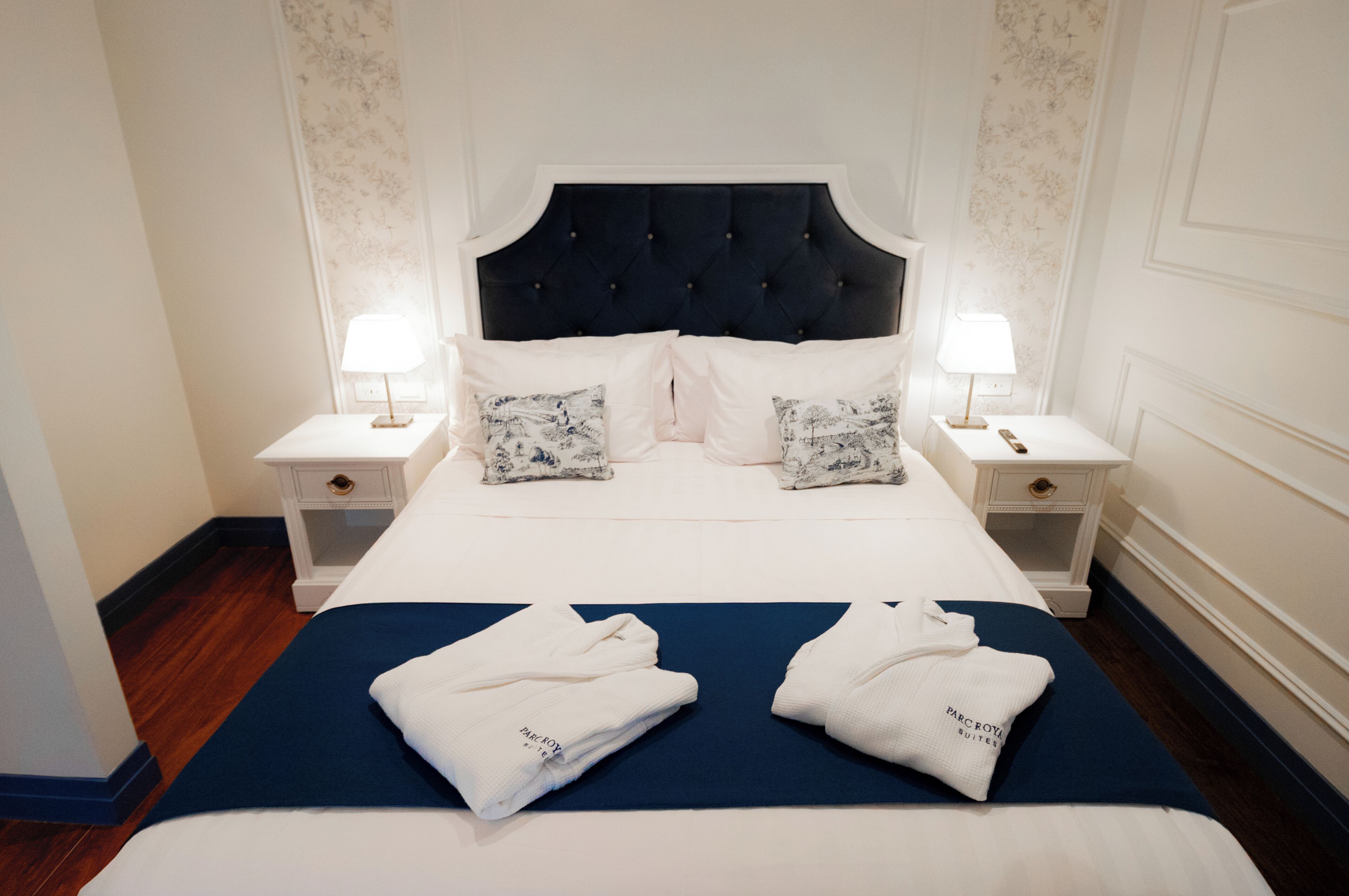 A hotel room with a large bed, two bedside tables with lamps, floral wallpaper, and a blue bedspread.