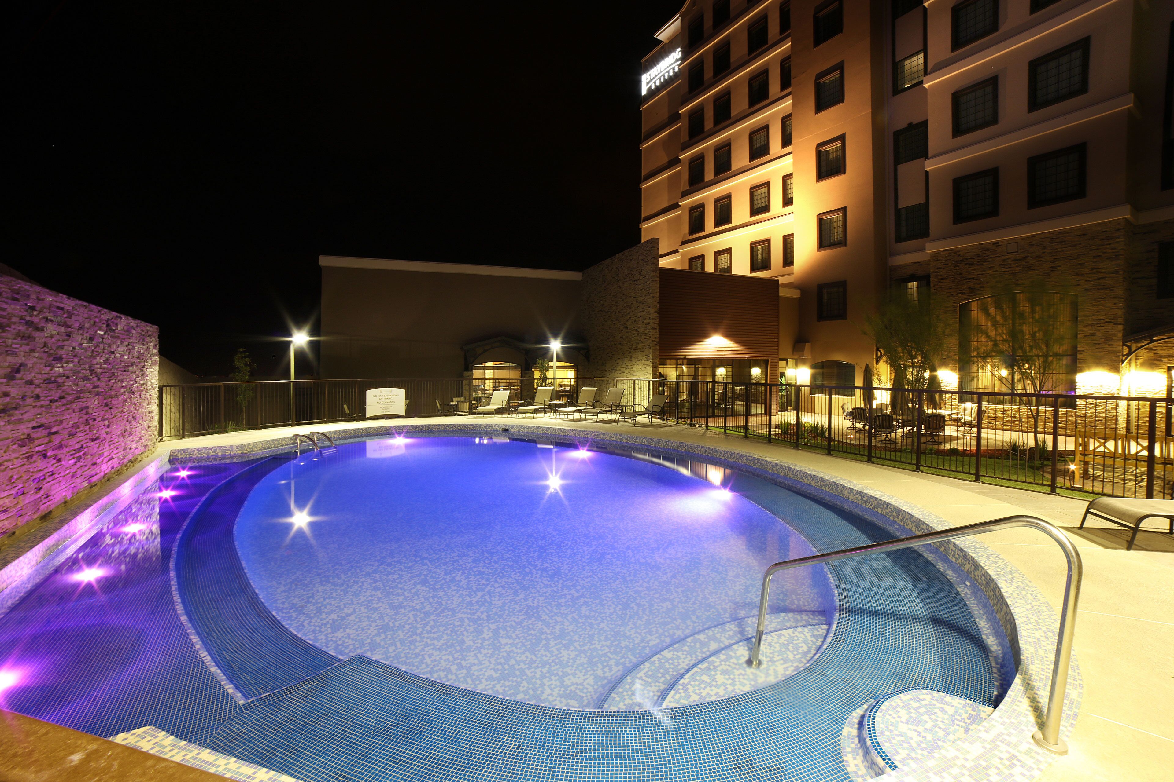 A swimming pool with a tiled edge, surrounded by a fence, and adjacent to a multi-story building with illuminated windows.
