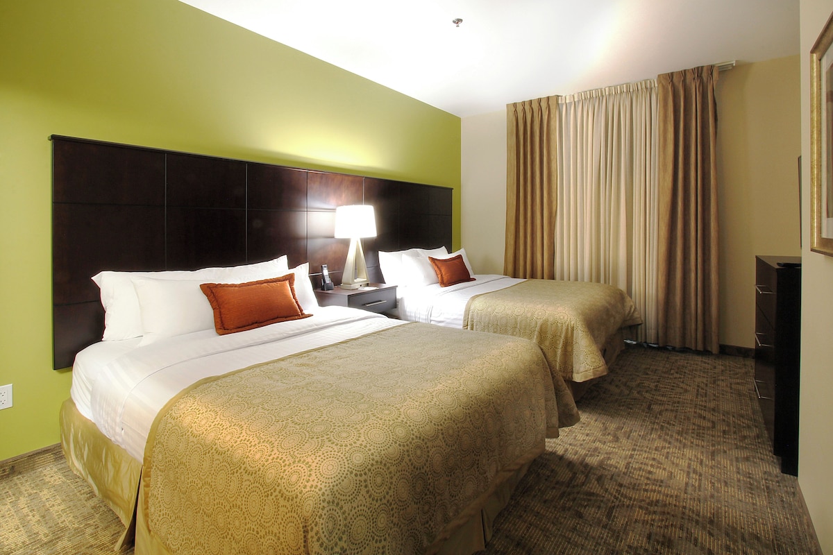 Een hotelkamer met twee bedden, een nachtkastje, een lamp en een groot raam met gordijnen.
