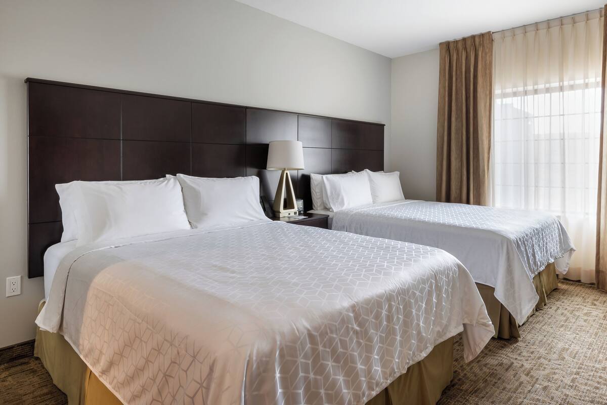 Een hotelkamer met twee bedden, een nachtkastje, een lamp en een groot raam met gordijnen.