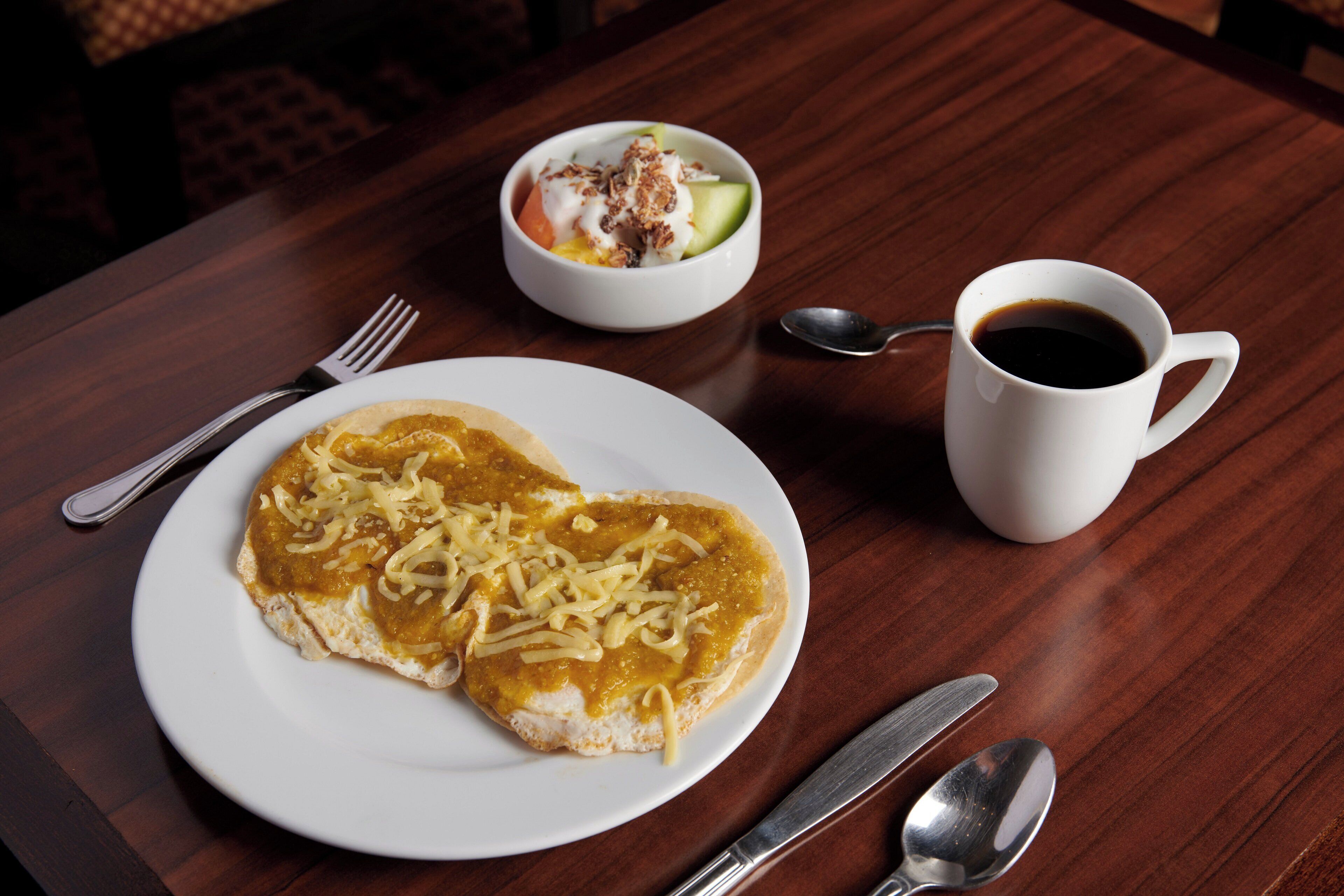 A plate of enchiladas with cheese, a bowl of salad, a cup of coffee, and cutlery on a wooden table.