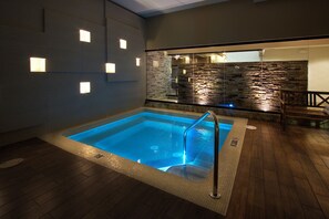 A modern indoor swimming pool with a glass barrier, wooden flooring, and stone wall backdrop.