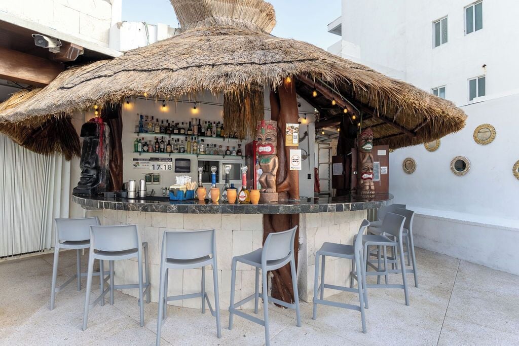 Eine Bar mit reetgedecktem Dach, Barhocker und einer Auswahl an Flaschen und Gläsern.