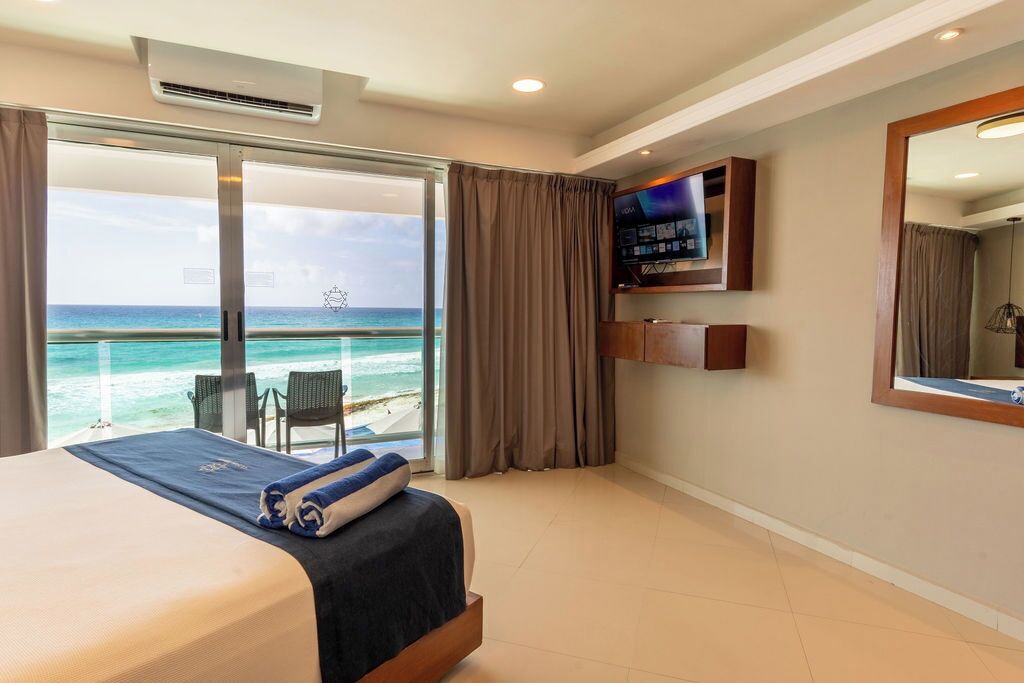 A hotel room with a bed, a TV, a large window with a view of the ocean, and a balcony with chairs.