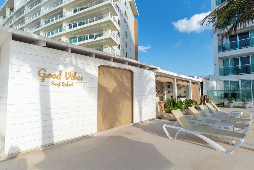 A building with 'Good Vibes Surf School' signage, a pool area with lounge chairs, and a clear blue sky.