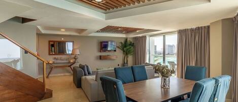 Un salon moderne avec un plafond en bois, un coin repas avec des chaises recouvertes d’un tissu bleu, un téléviseur et une grande fenêtre avec des rideaux.