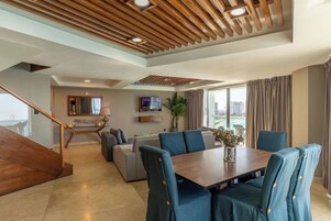 Un soggiorno moderno con soffitto in legno, un'area pranzo con sedie rivestite in blu, una TV e un'ampia finestra con tende.
