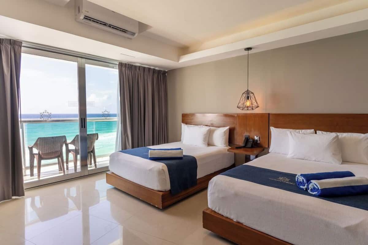 Ein Hotelzimmer mit zwei Betten, einem großen Fenster mit Meerblick, einem Deckenventilator und einem kleinen Tisch mit einer Lampe.
