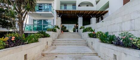 A modern building with a central staircase leading to an entrance, surrounded by lush greenery and a stone wall.