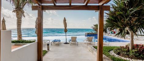 A covered outdoor area with a view of the ocean, lounge chairs, and a pool.