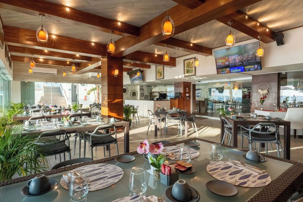 A restaurant with multiple dining areas, wooden beams, hanging pendant lights, and a television screen displaying a menu.