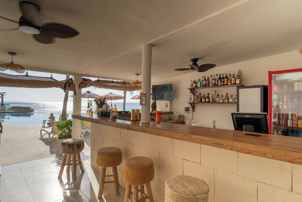 A bar area with a pool view, bar stools, ceiling fans, and a shelf with bottles.