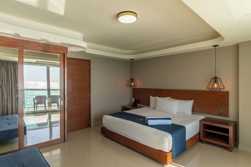 A hotel room with a bed, bedside tables, a nightstand, a chair, a large window, and a view of the sea.
