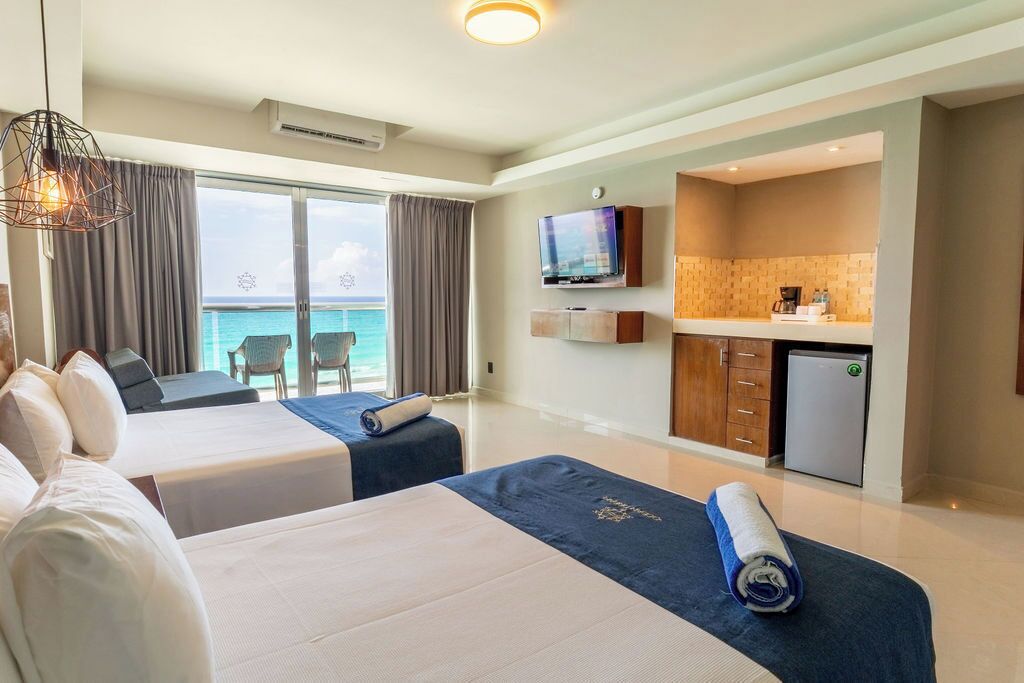 Quarto de hotel com duas camas, televisão, cozinha compacta e varanda com vista para o mar.