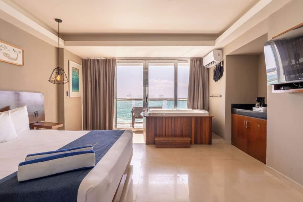 Ein Hotelzimmer mit einem großen Bett, einer kleinen Küchenzeile, einem Balkon mit Meerblick und einem an der Wand befestigten Fernseher.
