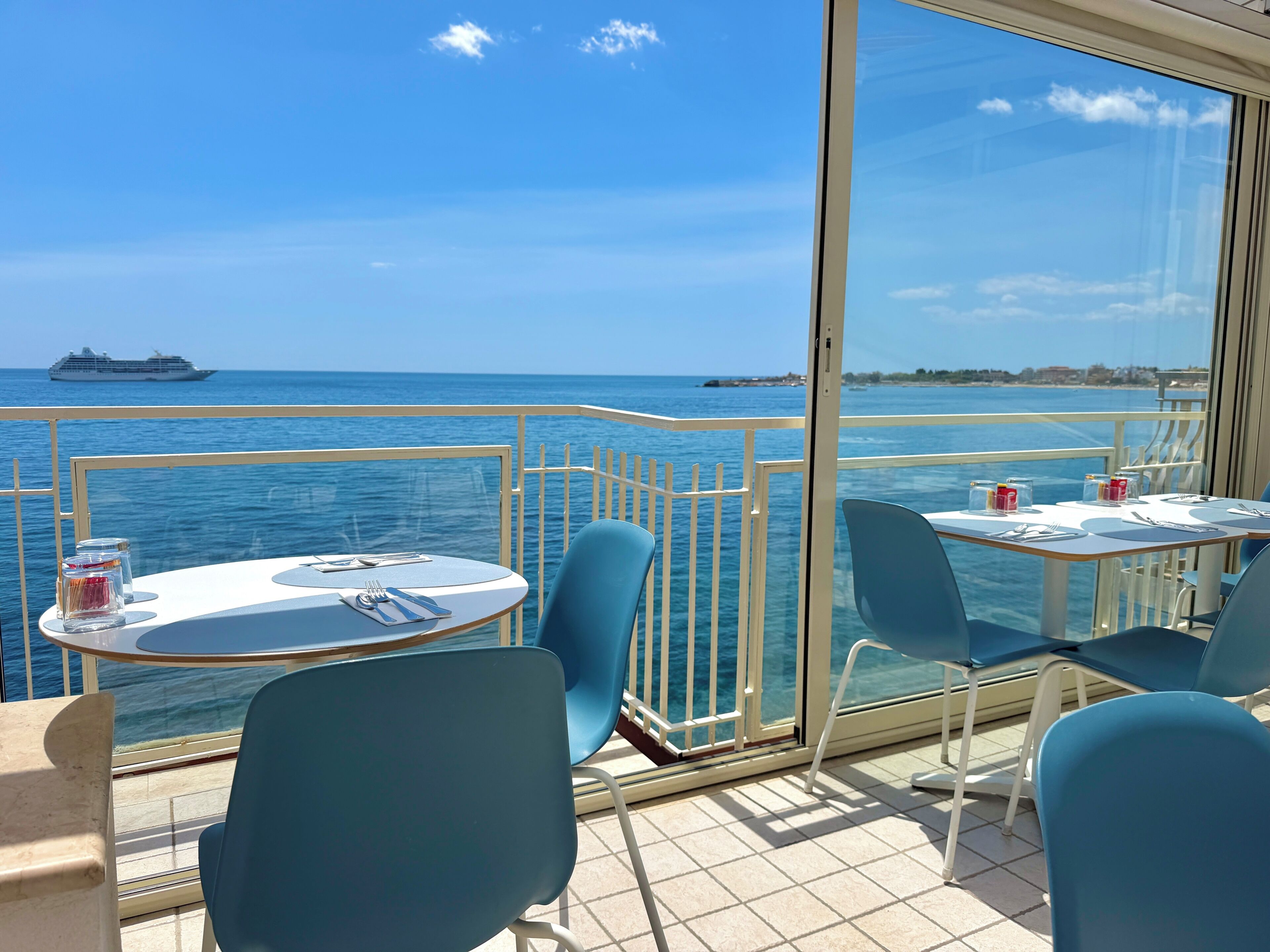 A balcony with tables and chairs, a ship in the background, and a clear blue sky.