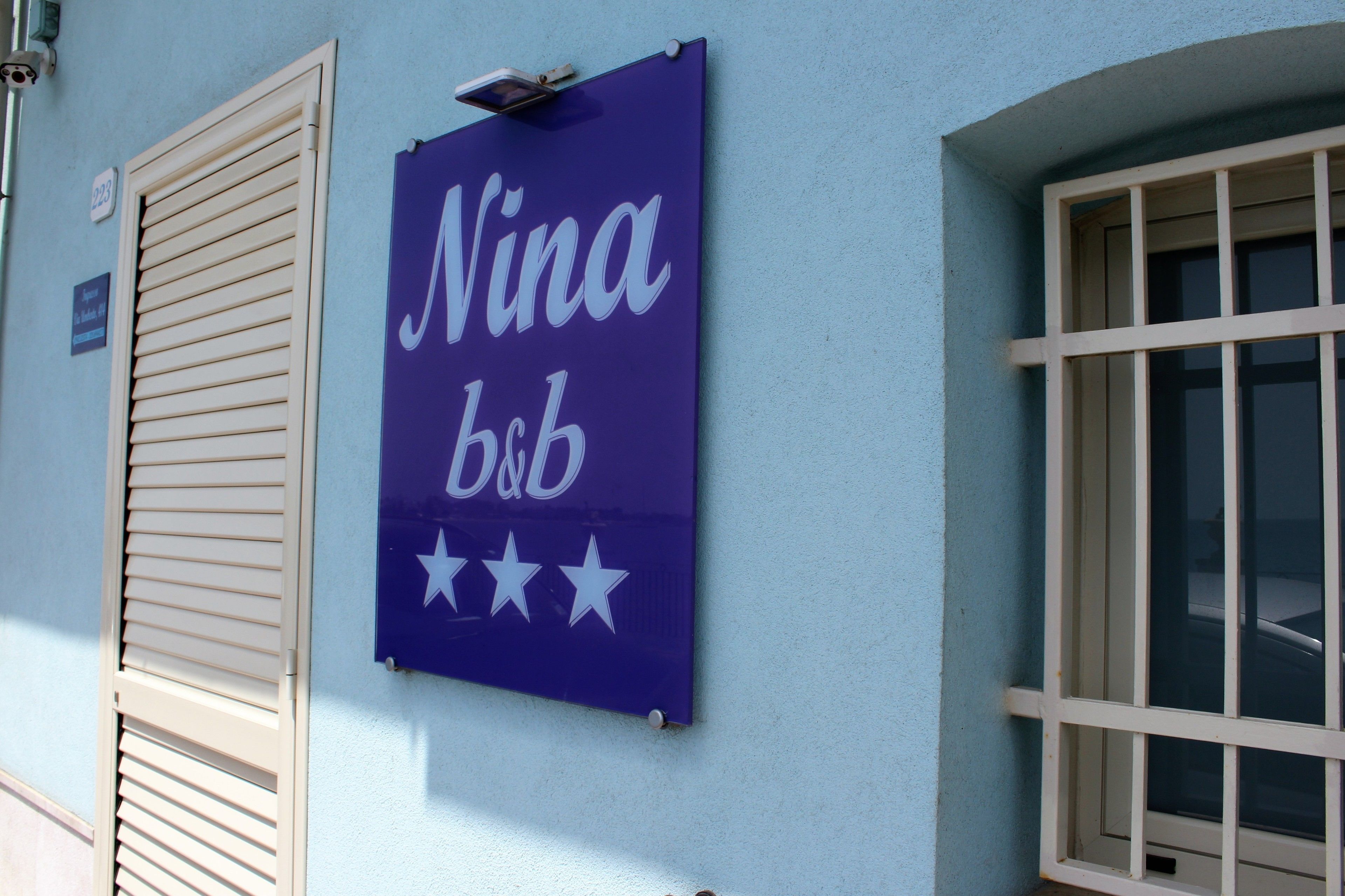 Un letrero que dice 'Nina b&b' sobre una pared azul claro, una ventana con persianas blancas, y un pequeño cartel encima de la ventana.