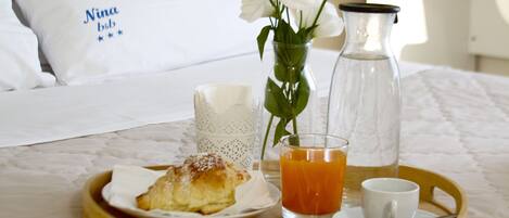 Ein Frühstücksbrett mit Gebäck, Orangensaft und einer Tasse auf einem Bett mit weißer Bettwäsche und einem Kissen mit Schriftzug.