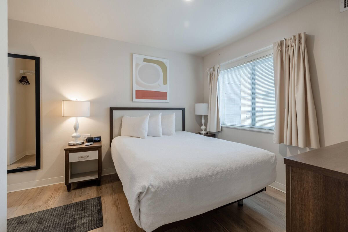 A neatly arranged hotel room with a bed, bedside tables, a mirror, and a window with curtains.