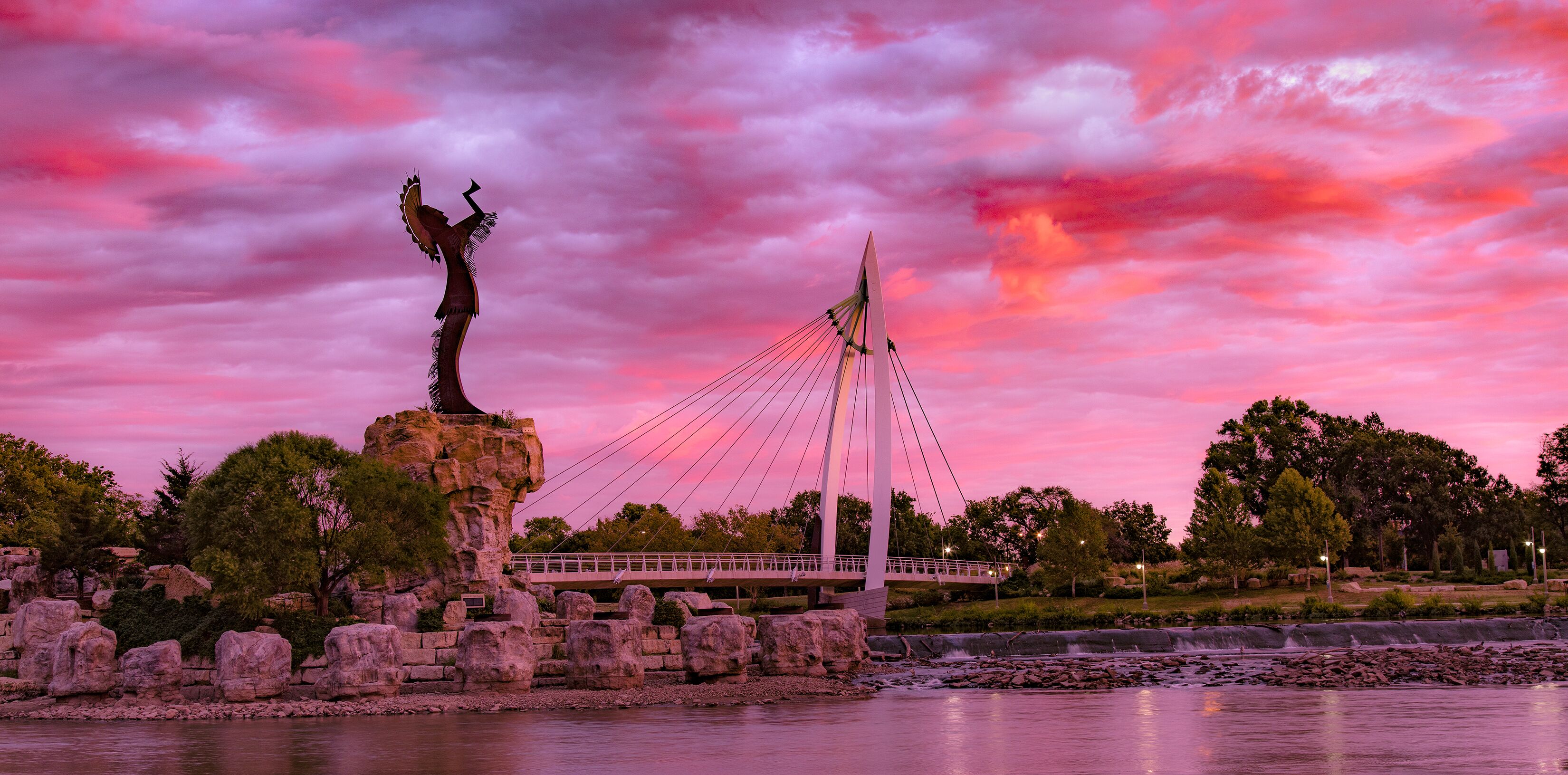 A suspension bridge with a statue on a rock, surrounded by trees and a body of water.