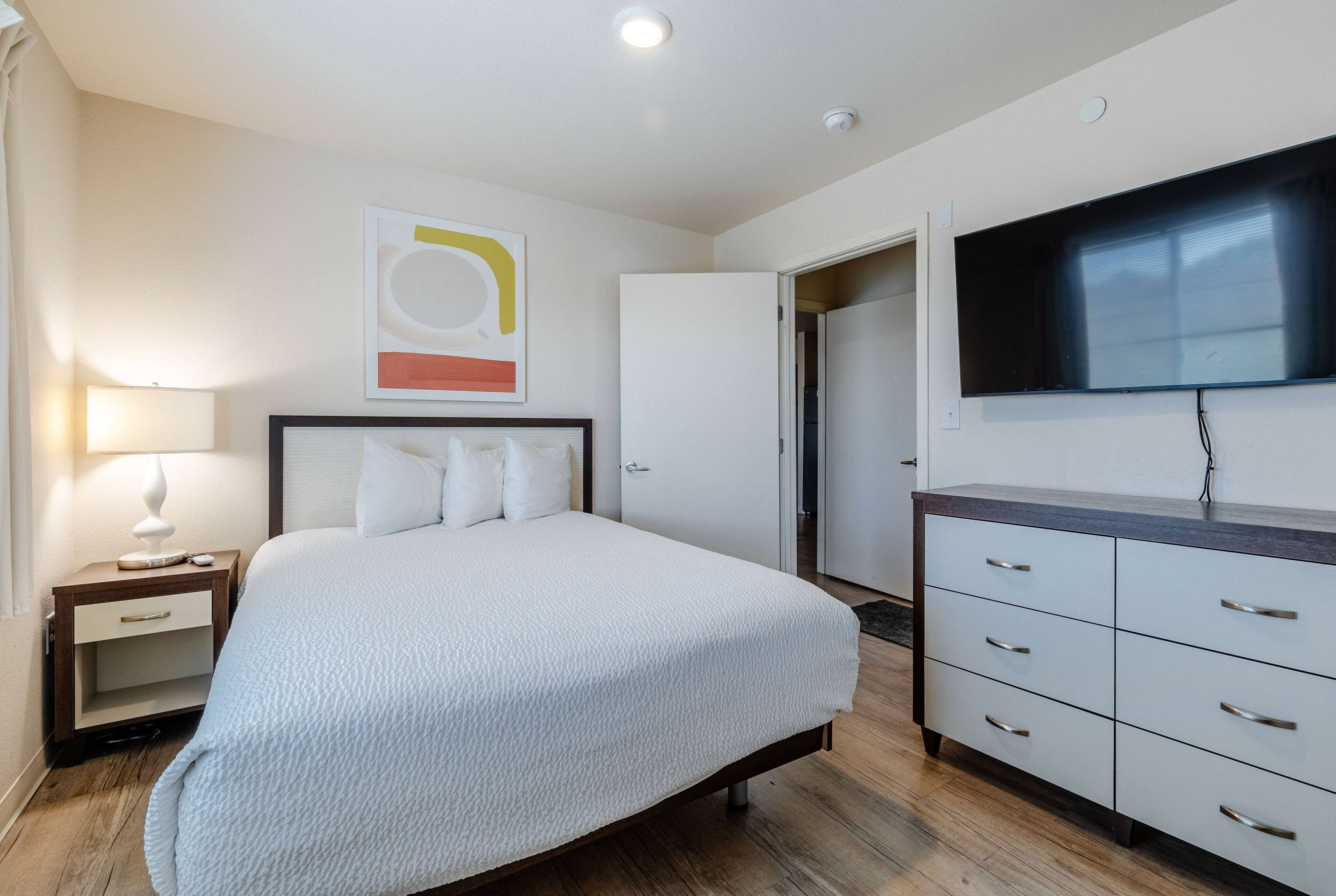 A bedroom with a bed, a nightstand, a dresser, a television, and a door leading to another room.