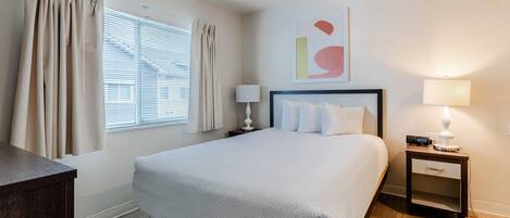 A hotel room with a bed, bedside tables, a lamp, a window with curtains, and a wall art piece.