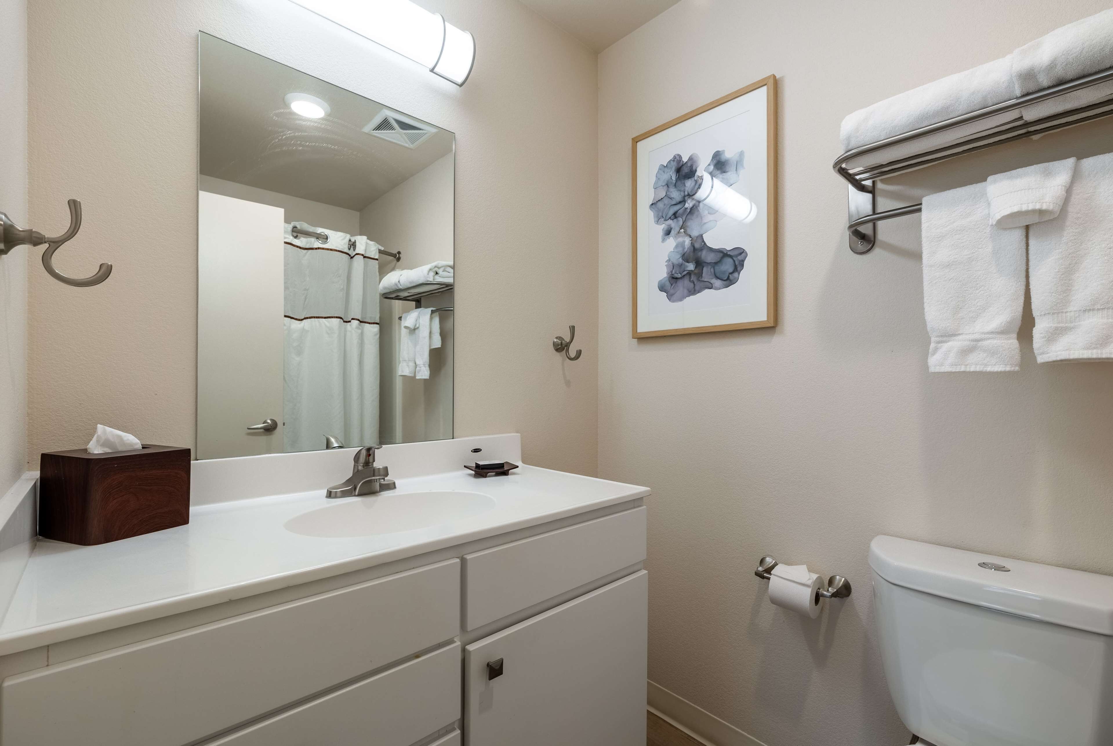 A bathroom with a white sink, mirror, and toilet. There is a towel rack with a towel on it, and a framed picture on the wall.
