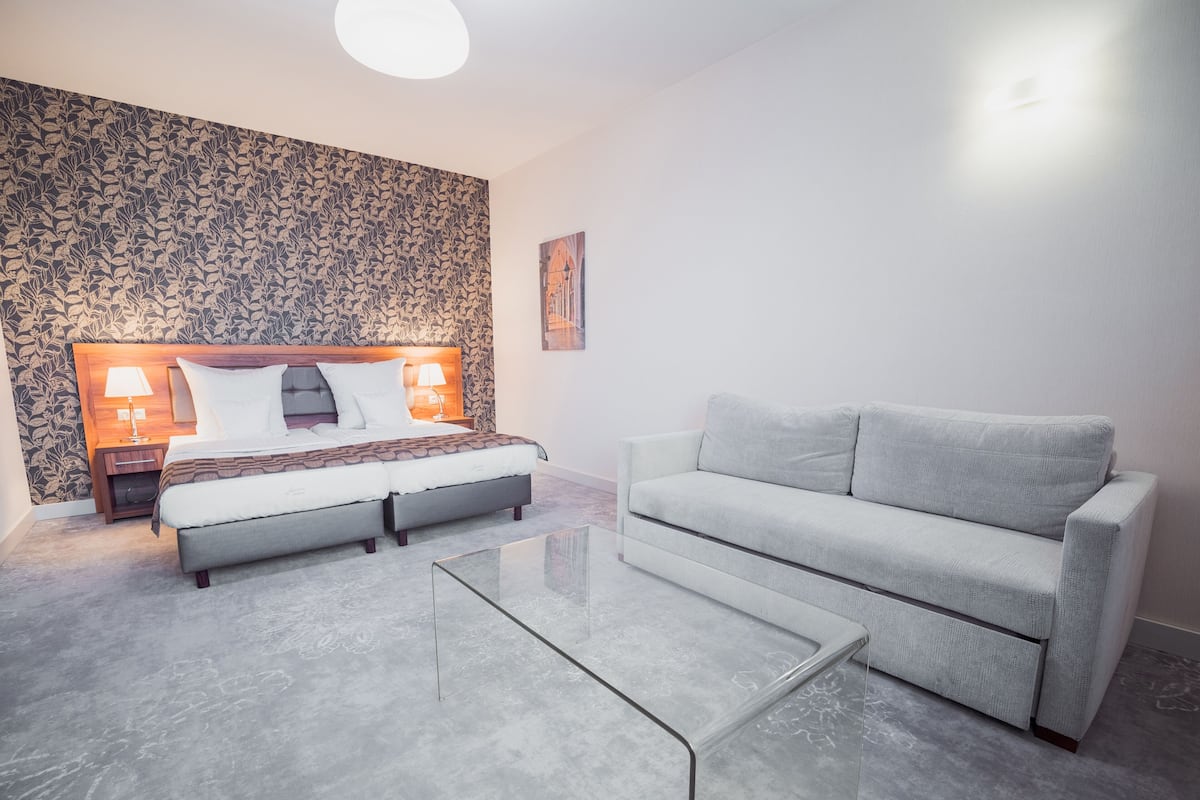 Quarto moderno com cama, sofá, mesa de centro de vidro e papel de parede estampado.